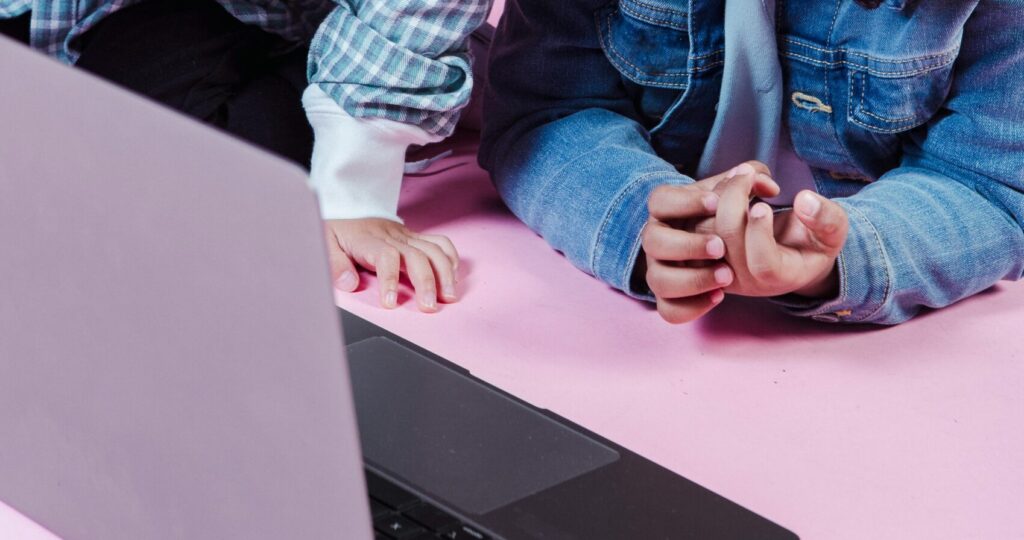 Multiracial children in casual clothes spending time in studio and sharing netbook on pink background