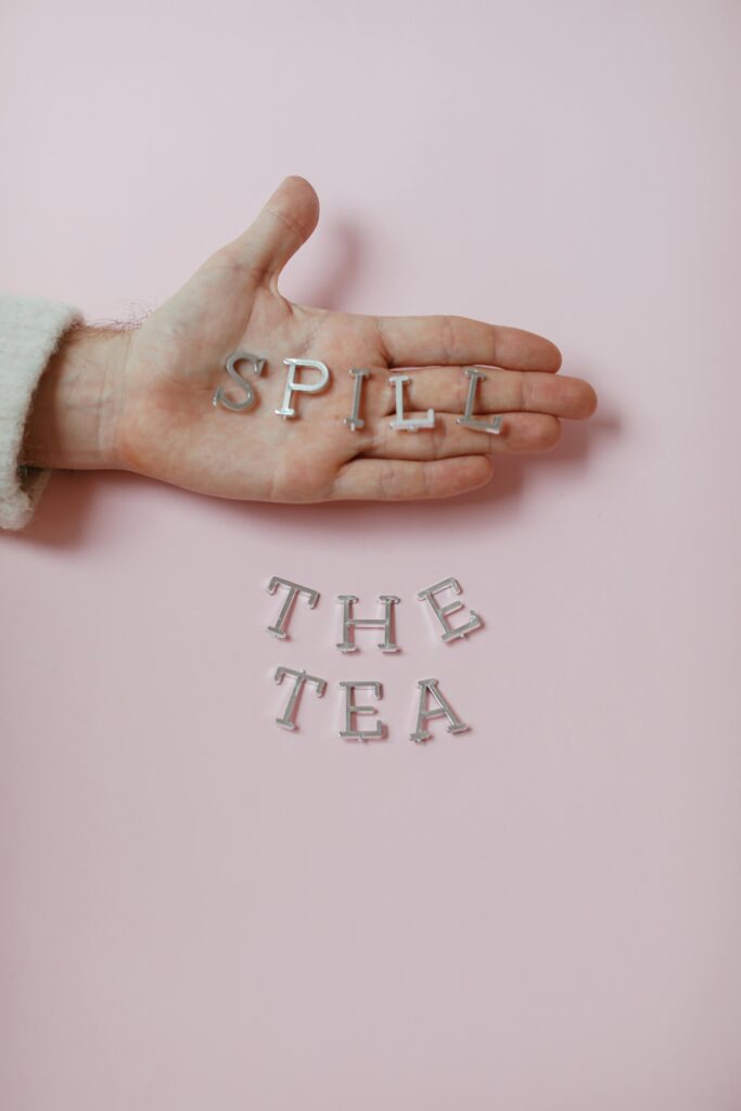 Hand holding 'SPILL' letters with 'THE TEA' on pink backdrop.