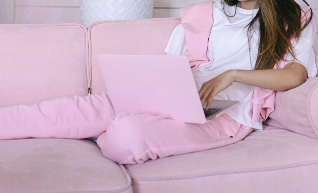 Young woman in pink outfit working on a laptop, relaxing on a cozy sofa with green plants in the background.
