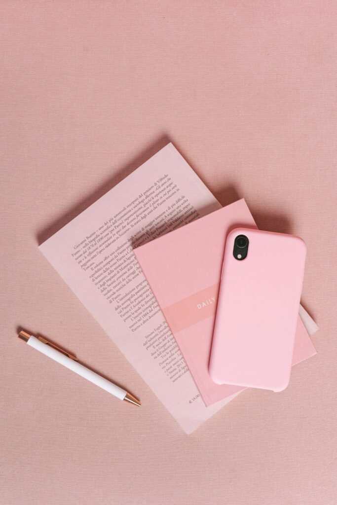 Flat lay of a pink themed workspace featuring a smartphone, notebook, and pen on a pink surface.